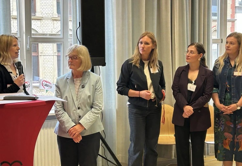 Fünf Frauen stehen in einem großen Raum an einem Rednerpult und hören einer Ansprache zu