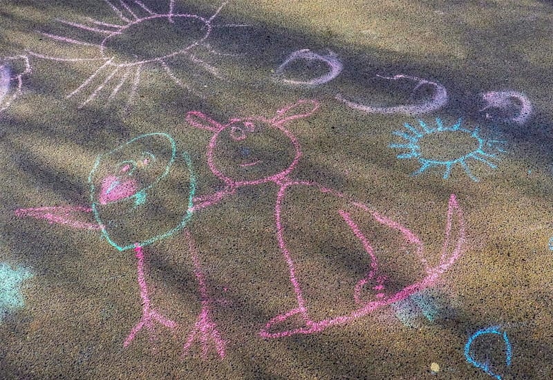 Farbenfrohe Kinderzeichnungen mit Straßenmalkreide auf grauem Asphalt, die eine Sonne und freundliche Figuren zeigen