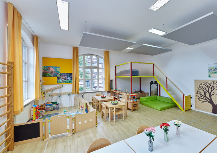 Heller Kita-Gruppenraum in Wiesbaden mit großer Spielhochebene aus Holz, Tischen und Stühlen für Kinder, Spielbereich mit Holzzaun und Sprossenwand. Im Hintergrund große Fenster und Schalldämmung an der Decke